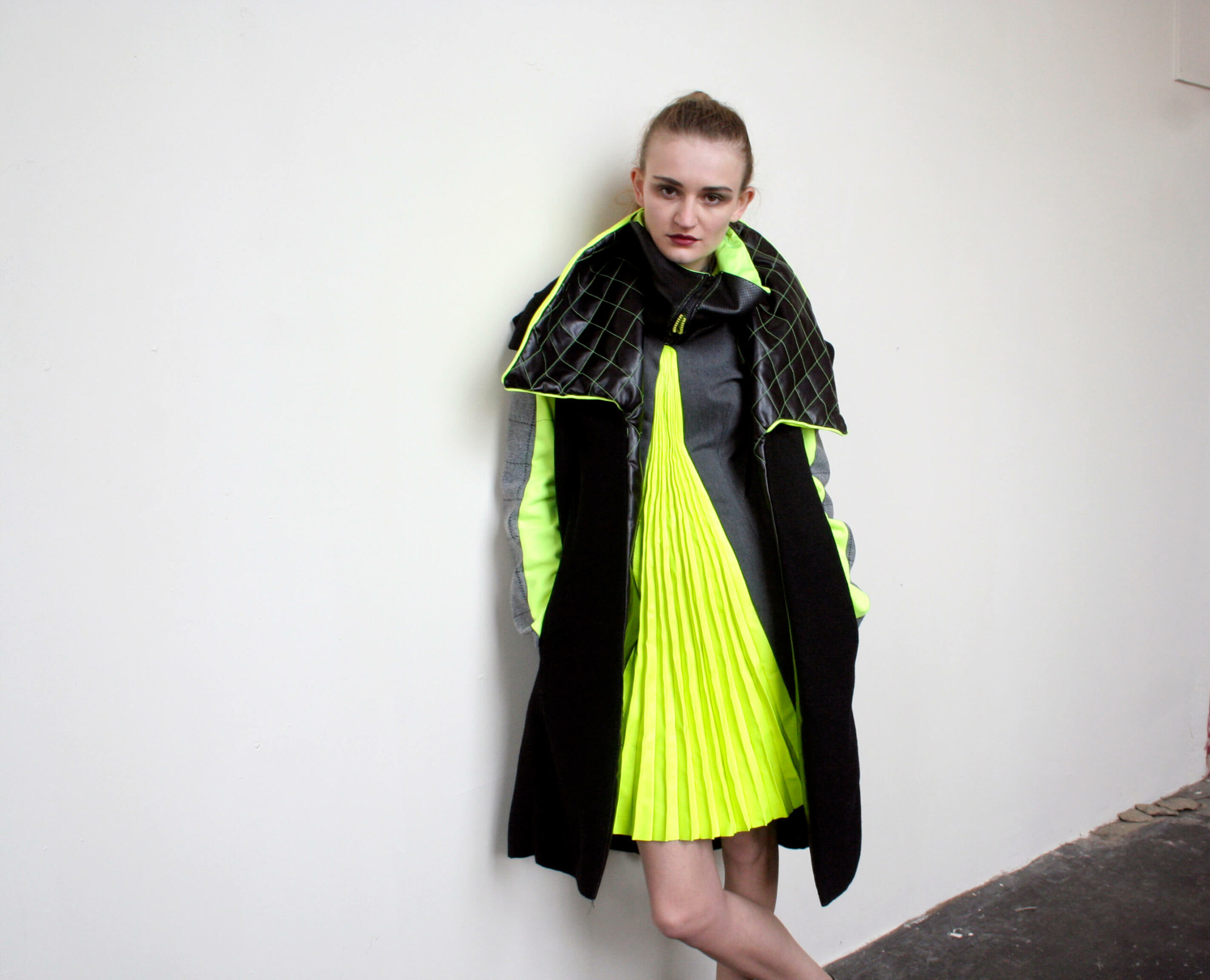 Photoshoot from the Armatura collection featuring a grey wool dress with neon green pleated leather insert and a black wool jacket with oversized quilted collar and neon stitching