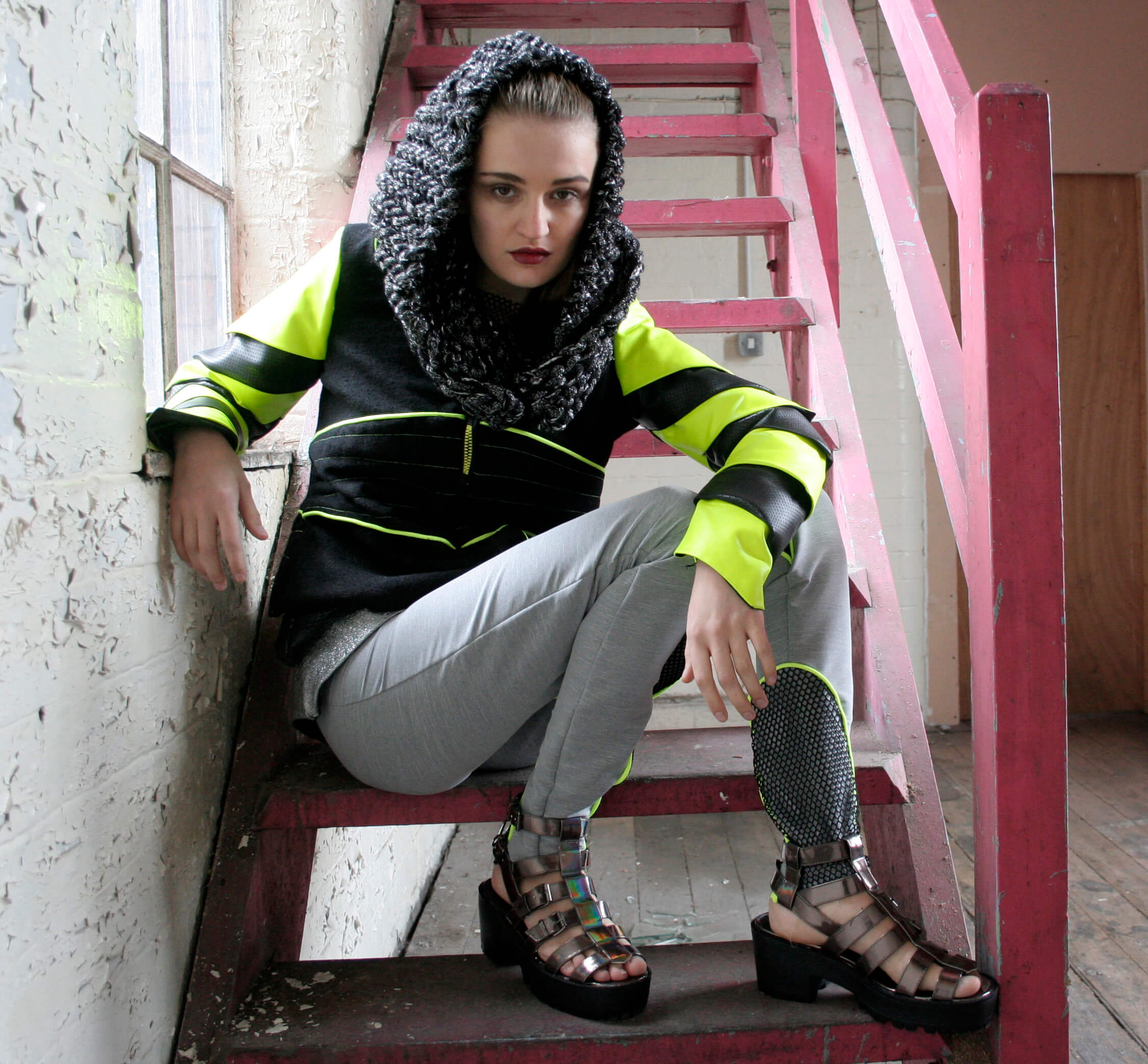 Photoshoot image from my university Armatura collection, featuring a model wearing a structured wool and neon jacket with a chunky knitted snood, grey stretch trousers with neon accents, and metallic gladiator sandals, seated on an industrial staircase